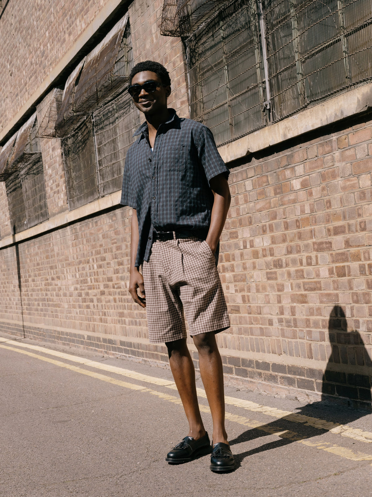Model wearing a dark check short sleeve shirt with checked tailored shorts and black leather loafers, photographed outdoors against an industrial brick wall.