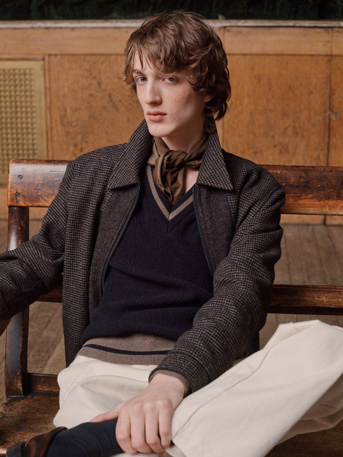 Man seated wearing a brown check jacket over a navy V neck jumper with a printed neckerchief