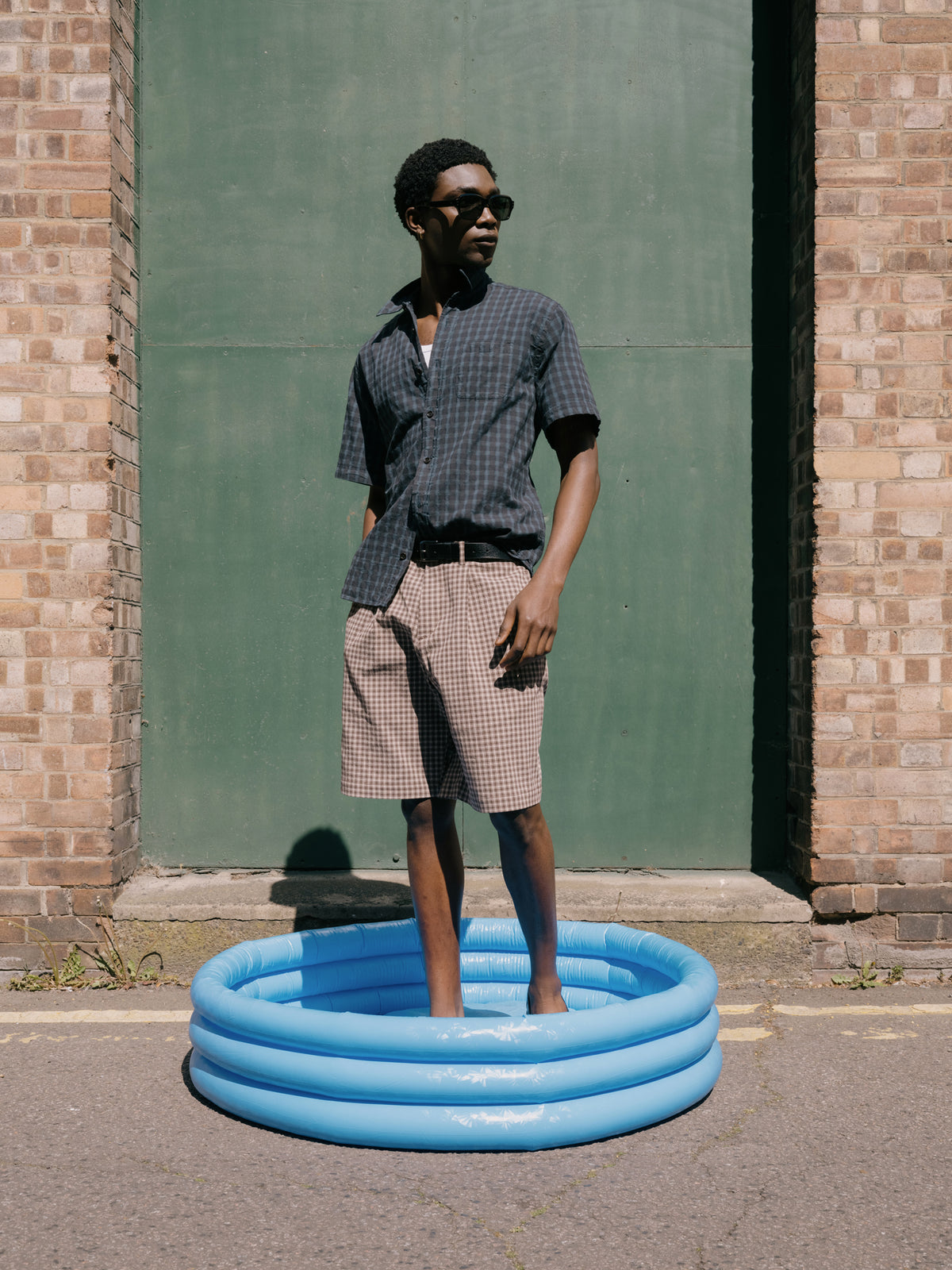 Navy check short sleeve shirt and brown check shorts, standing in a blue paddling pool against a green door.