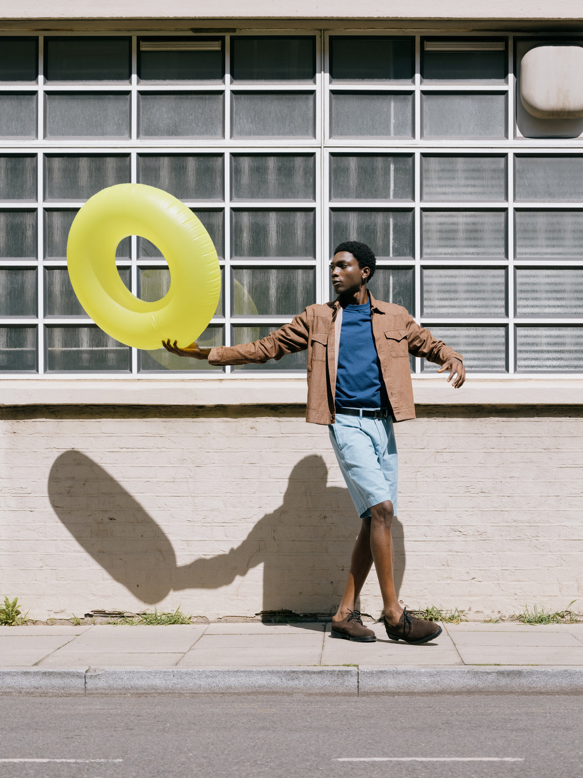 Tan Witley jacket worn open over a blue T-shirt with indigo pleated shorts, yellow ring on the wall behind.
