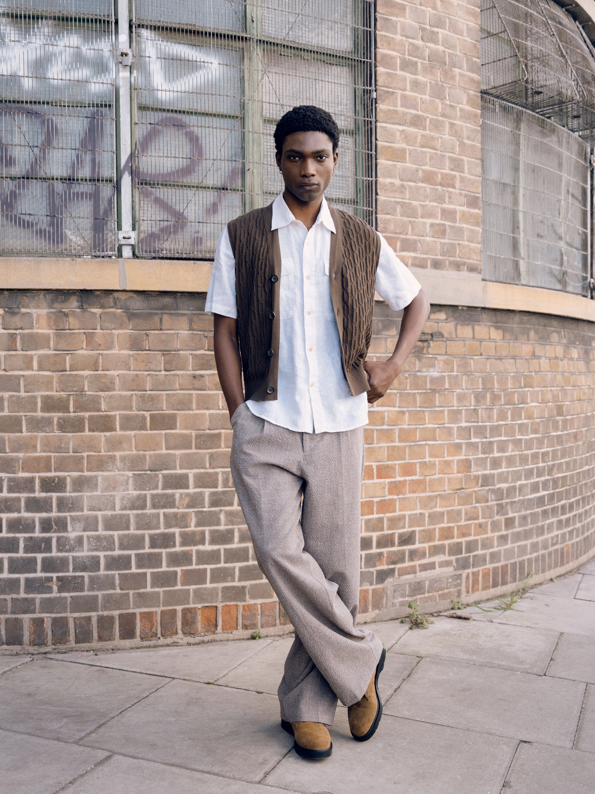 White short sleeve shirt under a brown knitted vest with grey pleated trousers, brick wall behind.