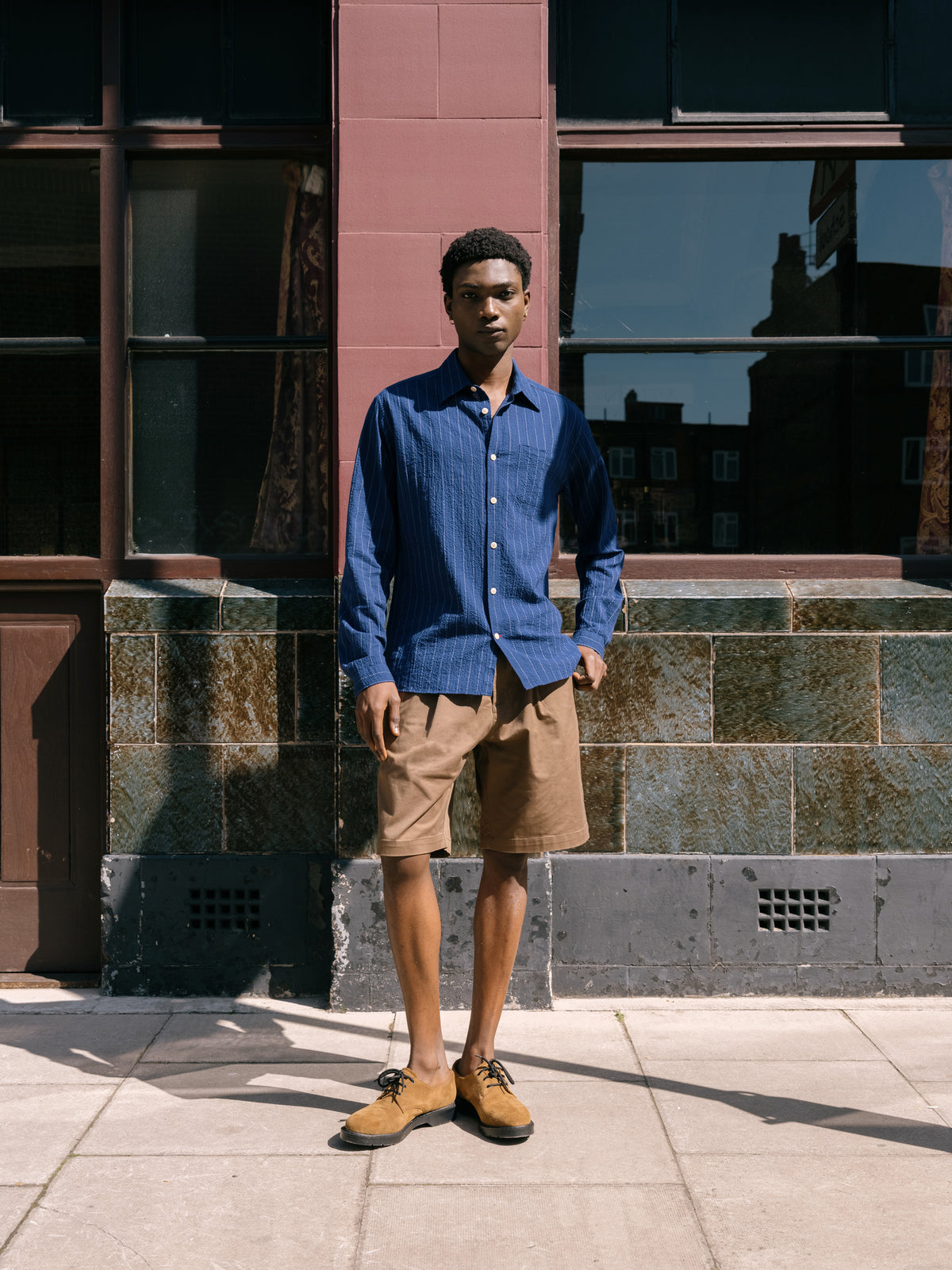 Blue long sleeve shirt and beige shorts with tan loafers, dark shopfront behind.