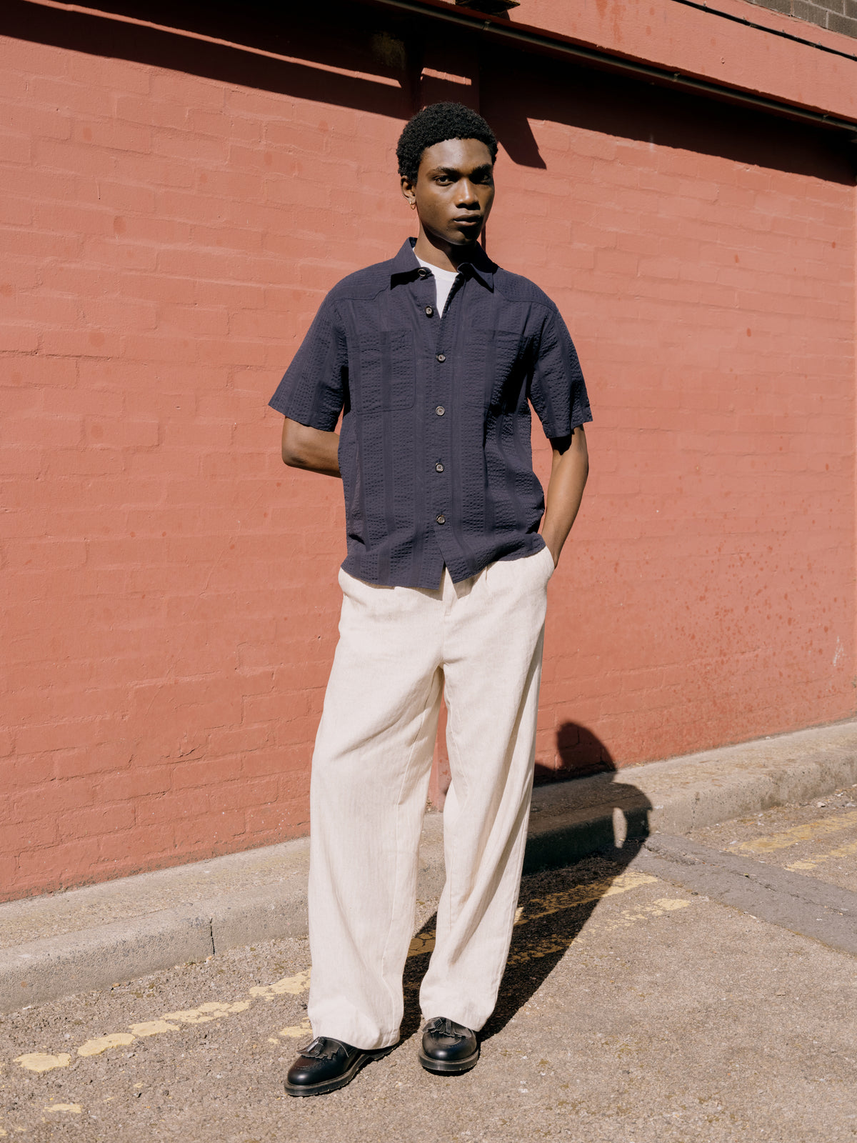 Navy short sleeve shirt over a white Oli's T-shirt with cream trousers against a red wall.