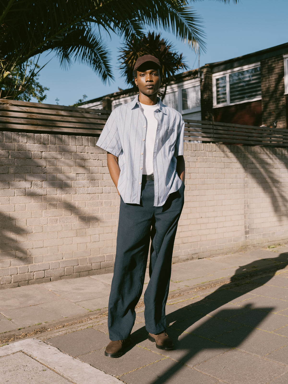 Pale striped short sleeve shirt with dark trousers, palm tree and painted wall behind.