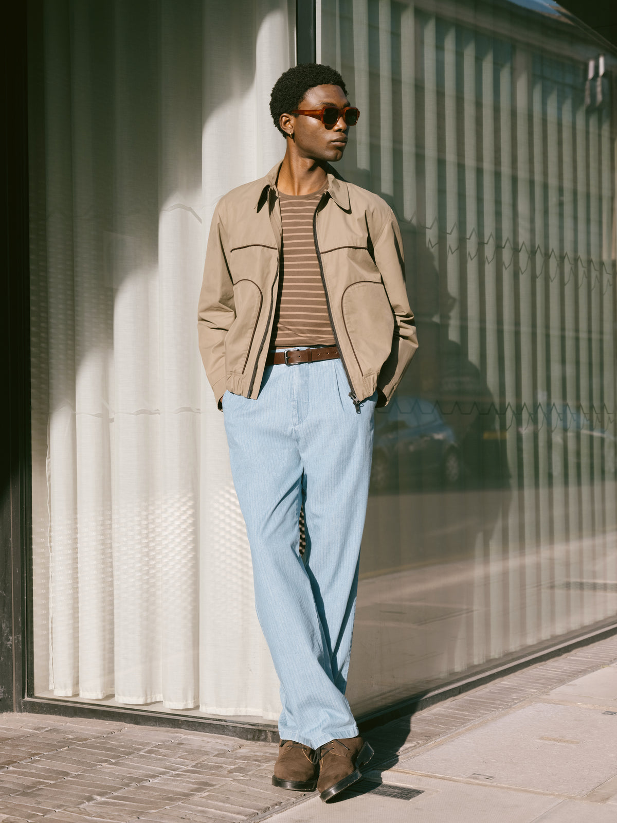 Khaki jacket over a striped T-shirt with pale blue trousers, ribbed metal wall behind.