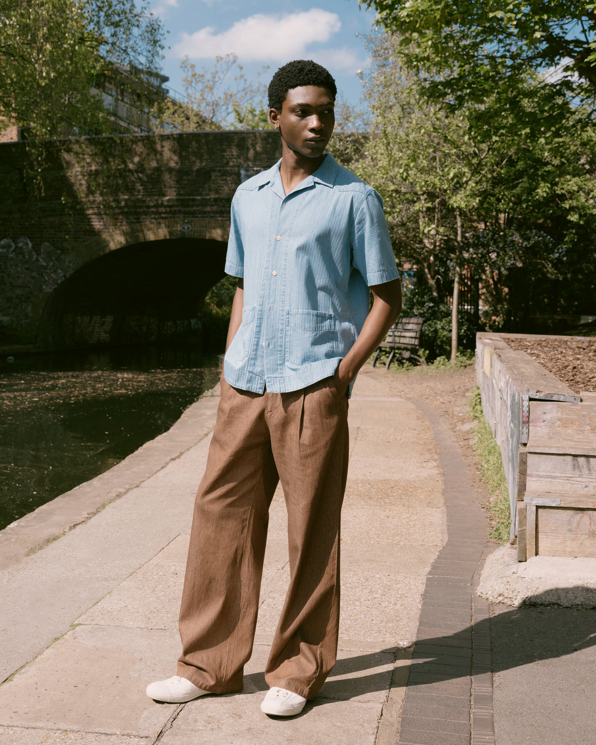 Indigo Cuban collar shirt with brown Orsman trousers on a towpath beneath a bridge.