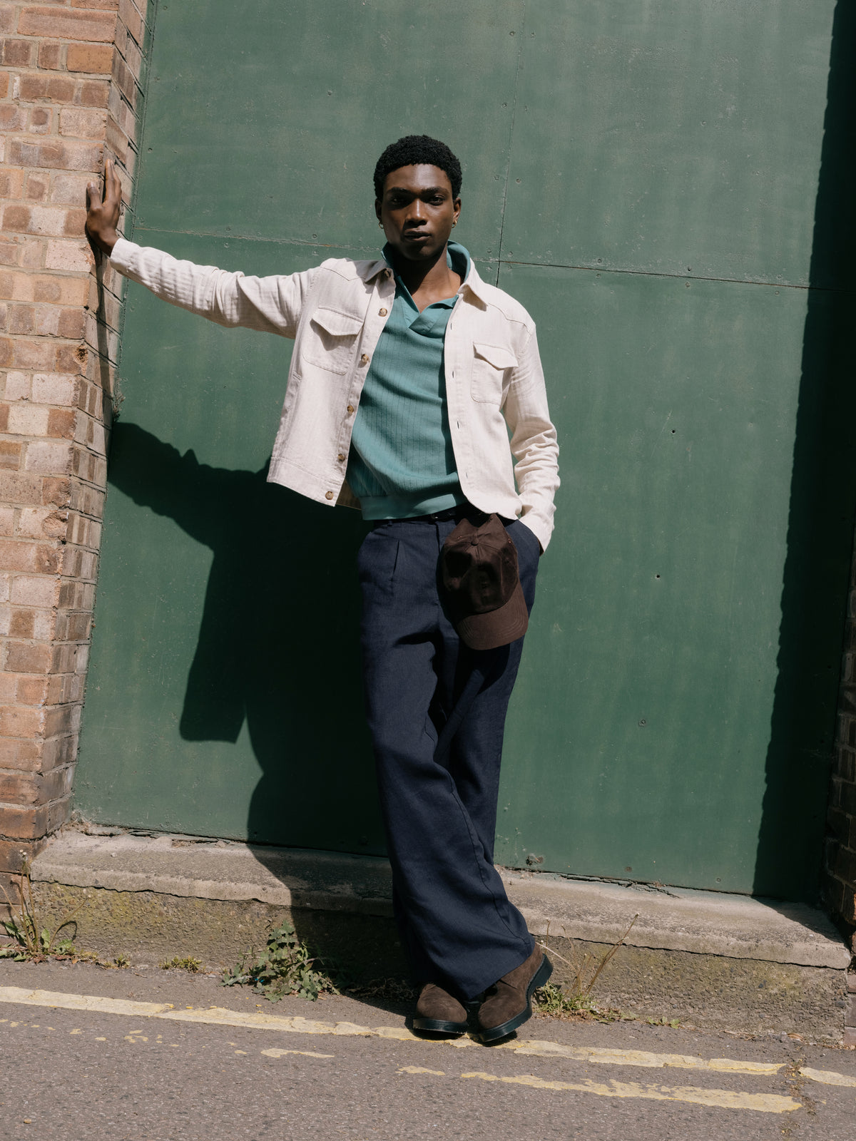 Cream jacket over a teal Westcott polo shirt with navy trousers, cap in hand against a green wall.