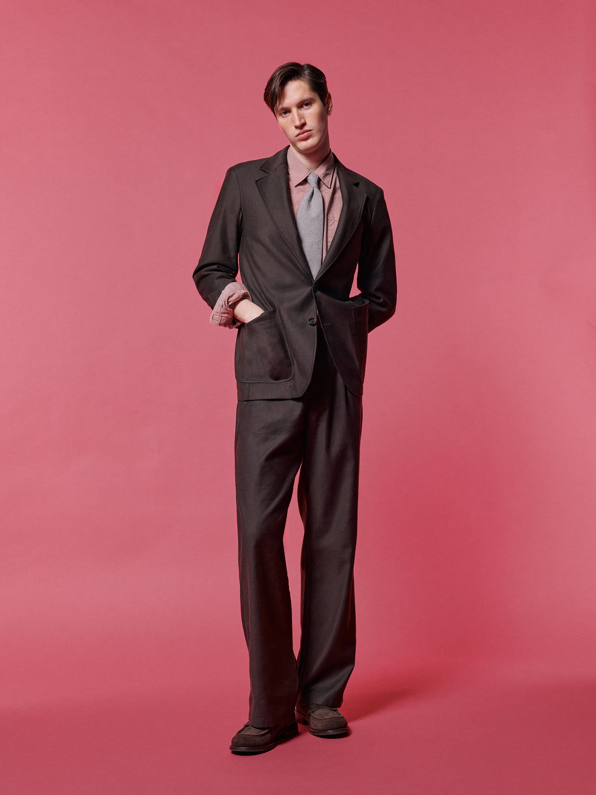 Brown suit with a tie, hands in pockets, against a pink backdrop.