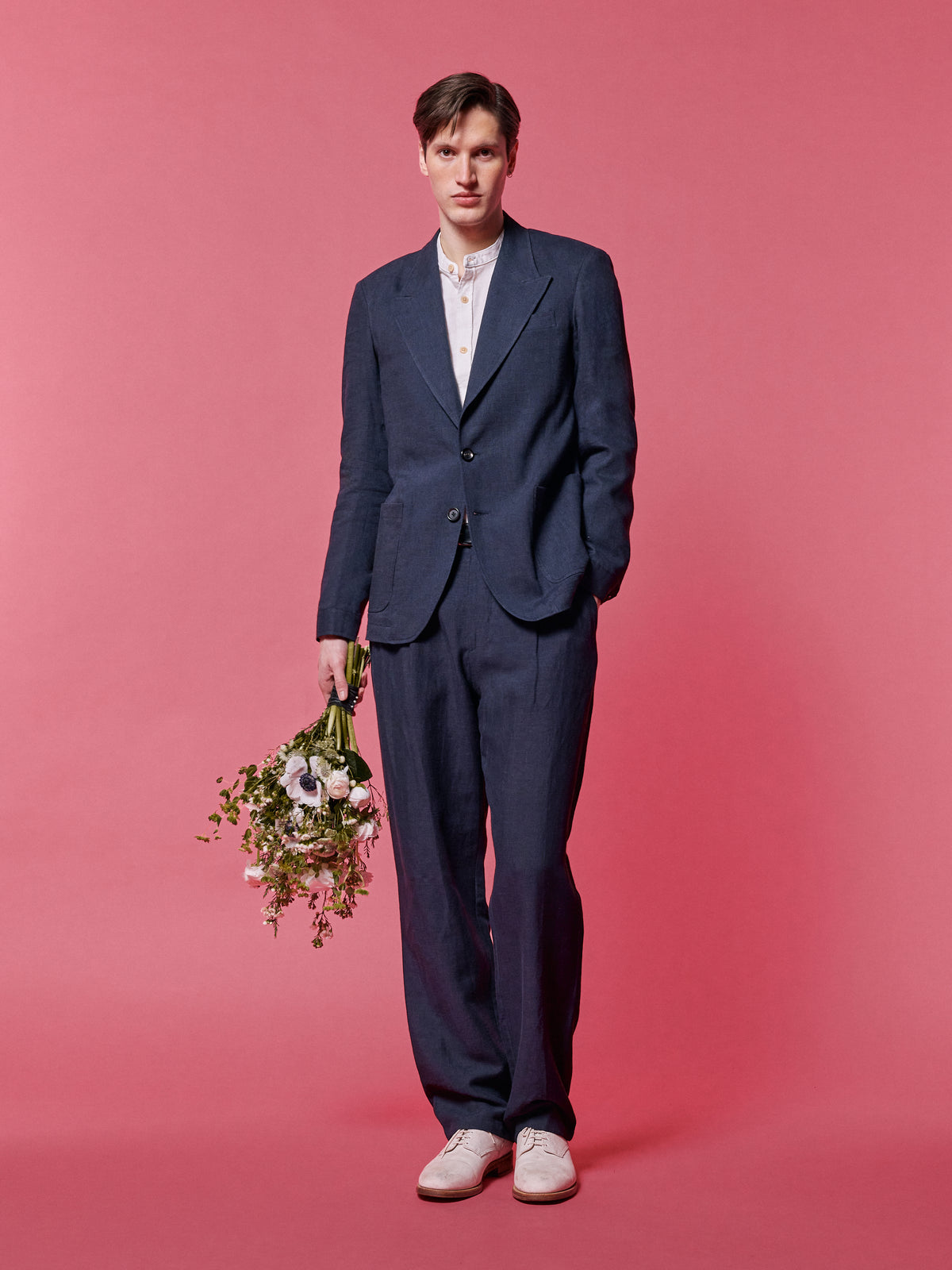 Navy suit with a grandad collar shirt, holding a bunch of flowers, against a pink backdrop.