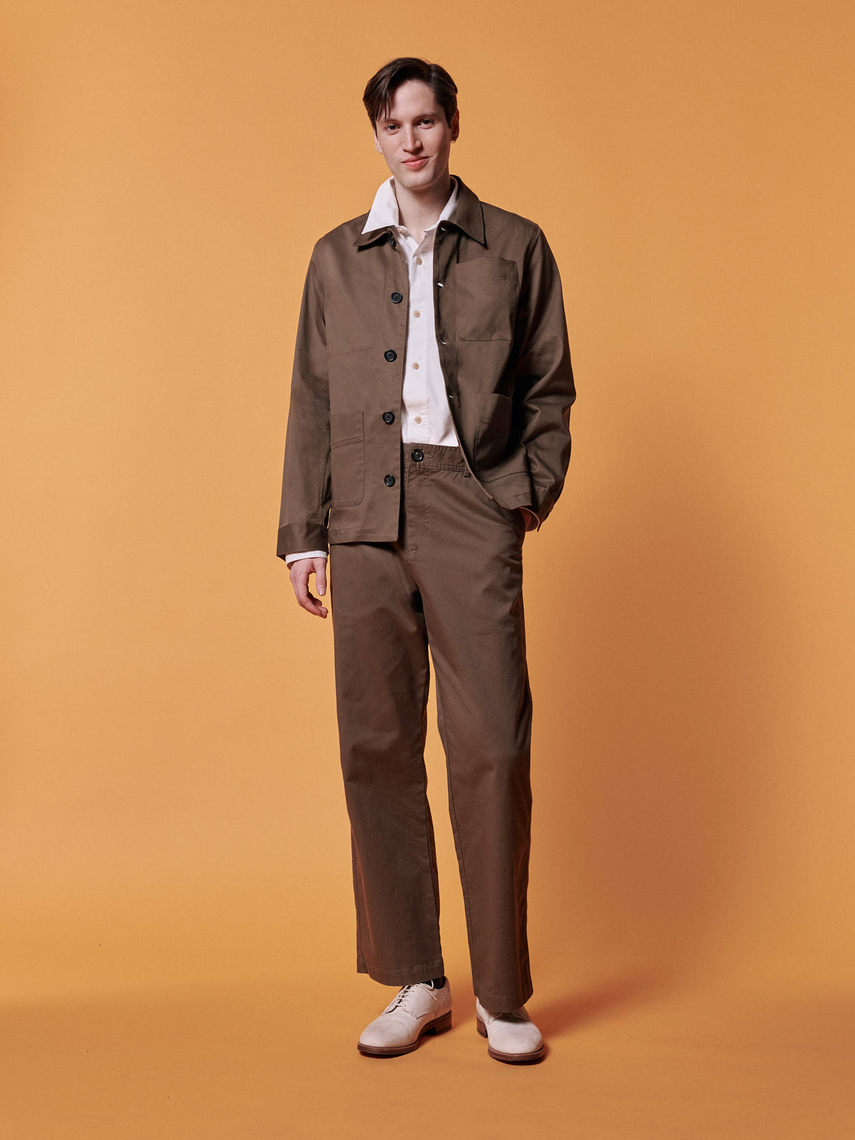 Brown jacket and matching trousers over a white shirt, against an ochre backdrop.