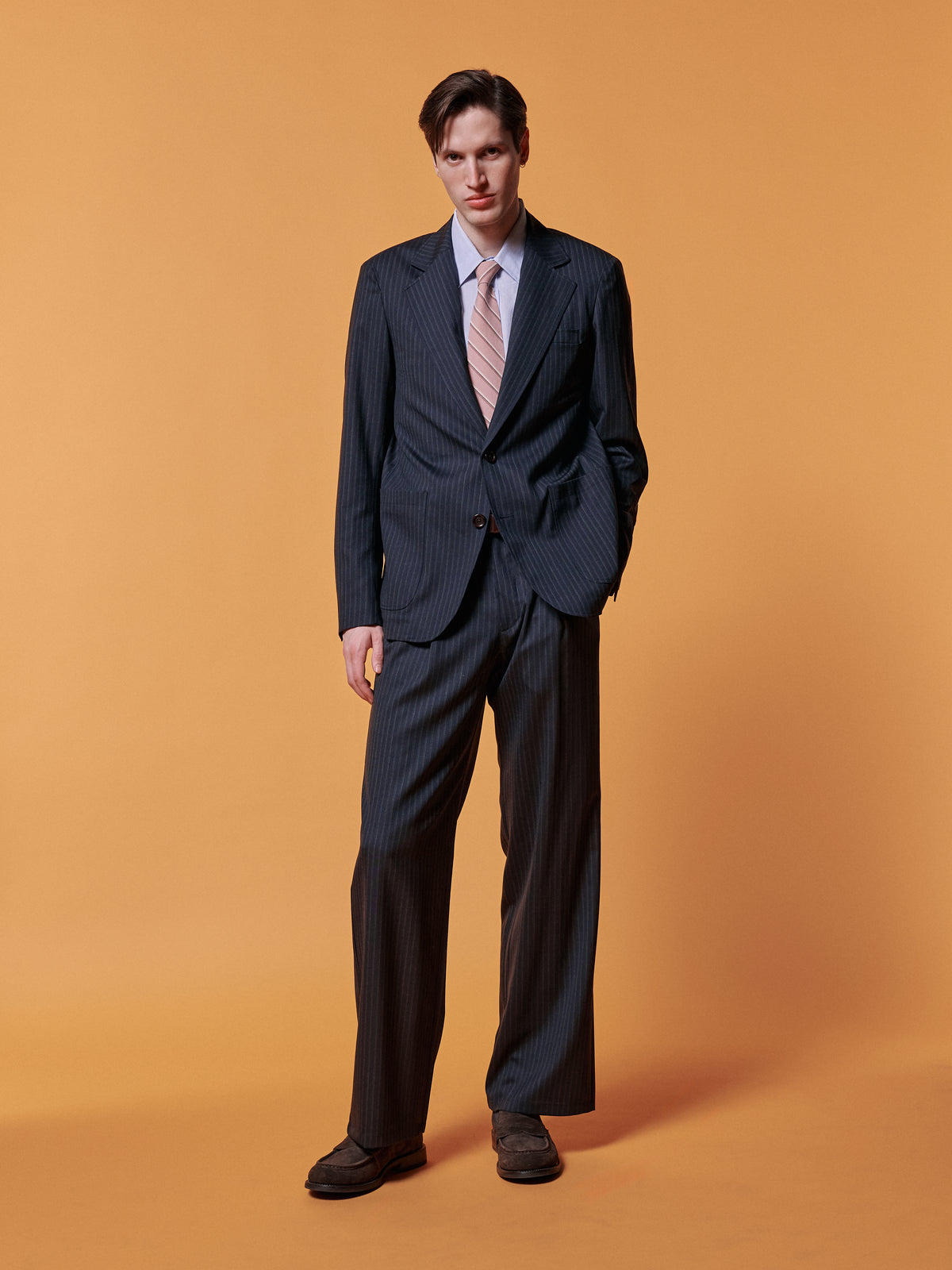 Navy suit with a blue shirt and pink tie, against an ochre backdrop.