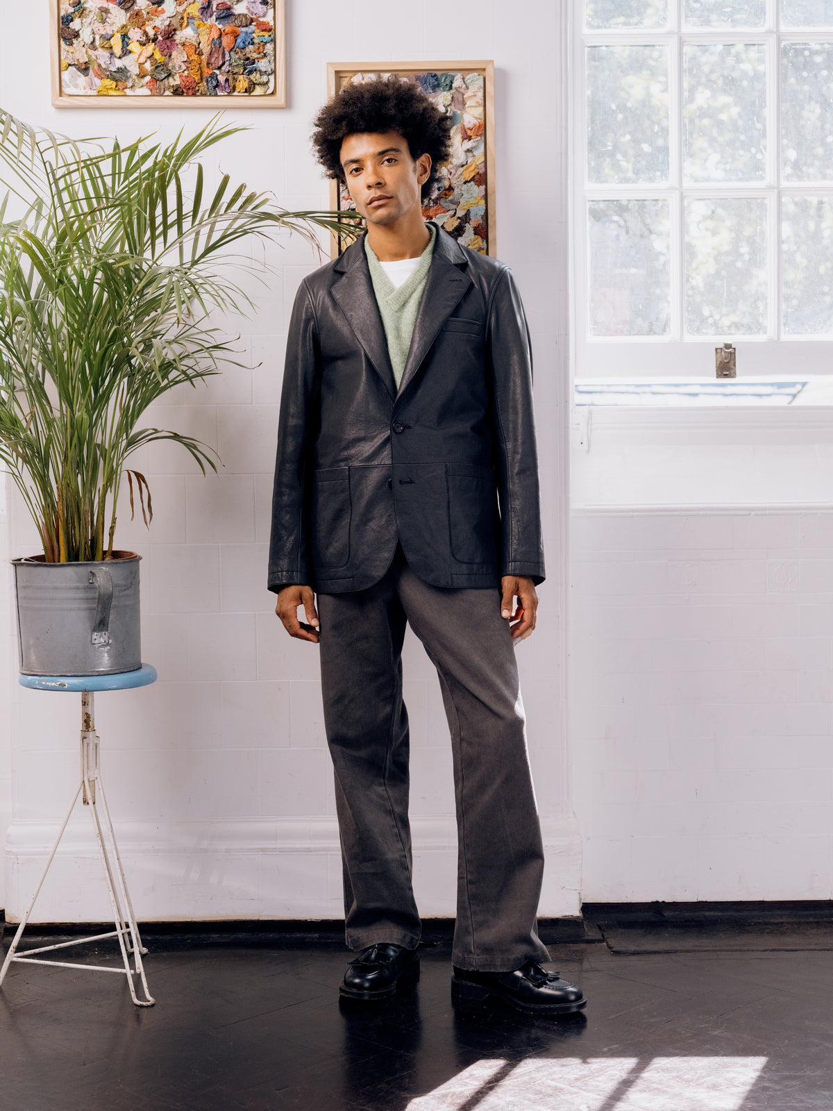 Black leather Westbury jacket over a pale green jumper with grey trousers, beside the potted palm.