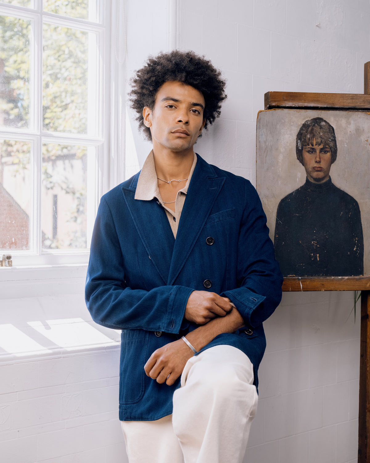 Indigo double breasted jacket over a pale shirt with ecru trousers, seated by the window.