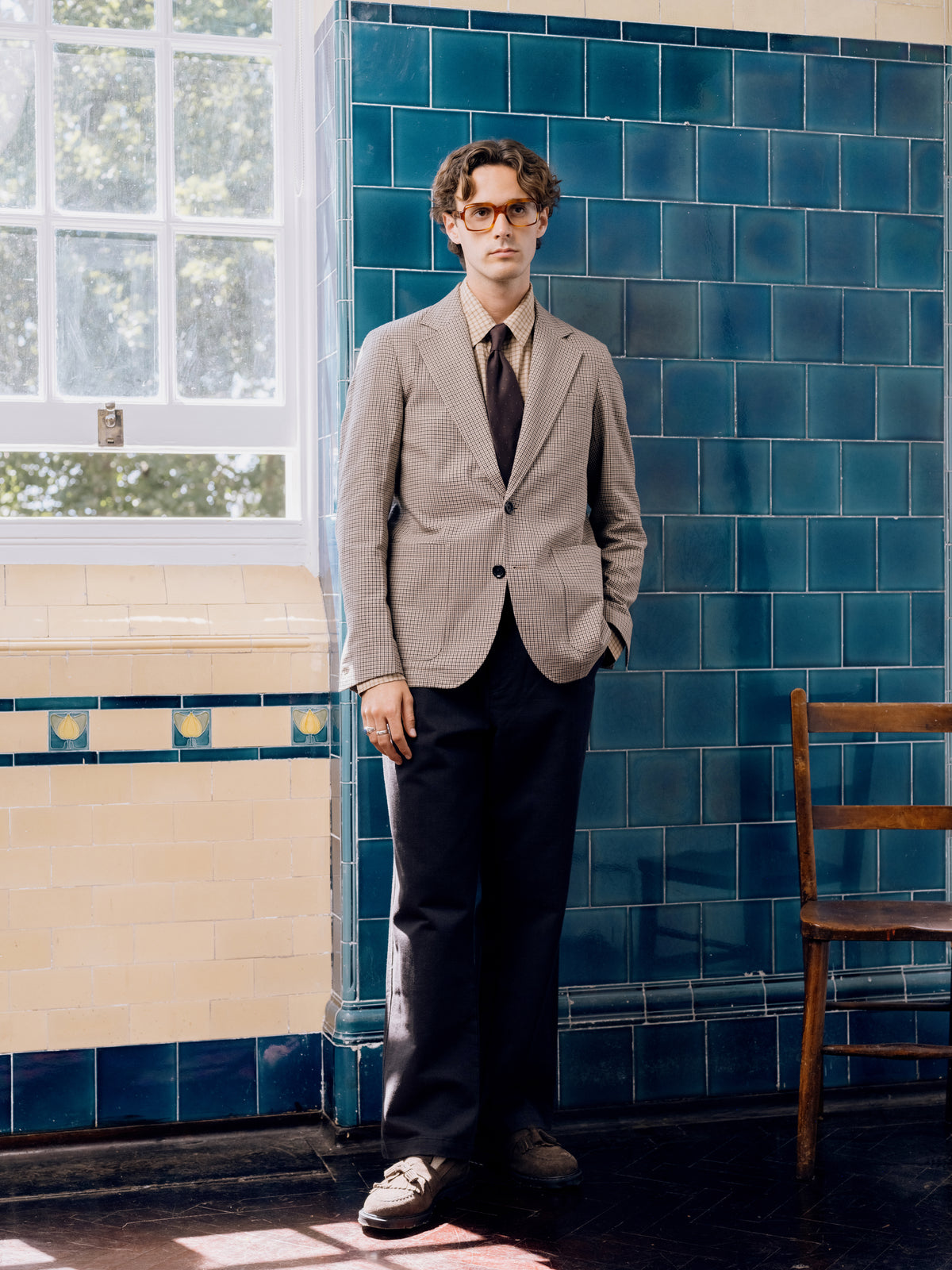 Houndstooth suit with a brown tie and tan suede shoes against the blue tiled wall.