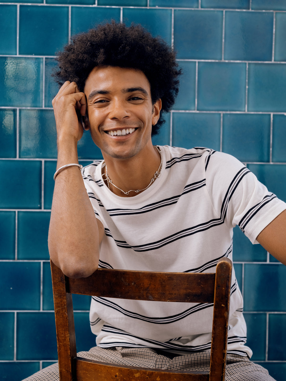 Cream and navy striped T-shirt with houndstooth trousers, seated on a wooden chair.