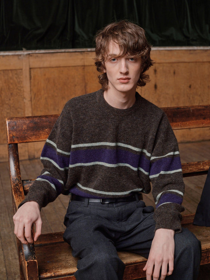 Man seated on a wooden bench wearing a brown and purple striped wool jumper with charcoal trousers