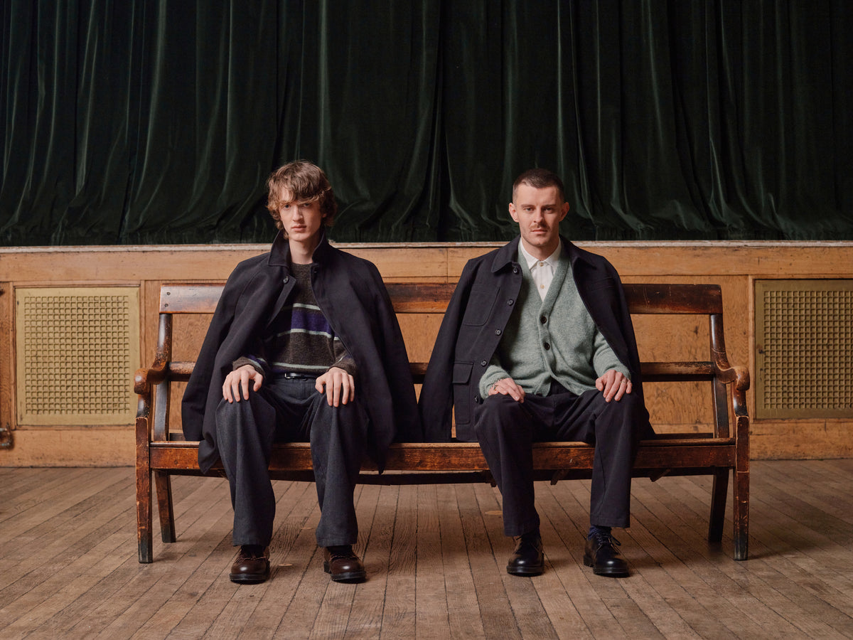 Two men seated on a wooden bench in a panelled hall, wearing Oliver Spencer Autumn 2026 coats, knitwear and tailored trousers