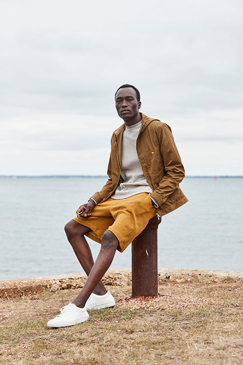 Oliver Spencer Spring 21, look 3. Seated on the beach in a tan jacket, ochre trousers and white trainers.