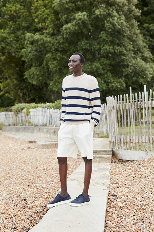 Oliver Spencer Spring 21, look 5. Breton striped jumper and white shorts on a path beside a fence.