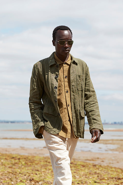 Oliver Spencer Spring 21, look 6. Olive overshirt and pale trousers on a shingle beach.