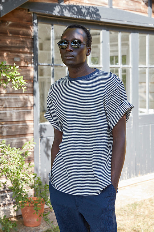Oliver Spencer Spring 21, look 12. Grey striped short sleeve knit and navy trousers beside a greenhouse.