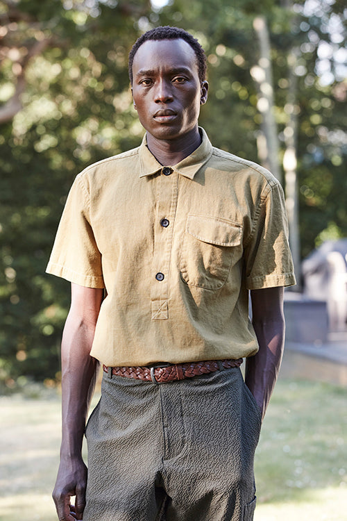 Oliver Spencer Spring 21, look 19. Mustard short sleeve shirt and olive trousers by a wooden cabin.