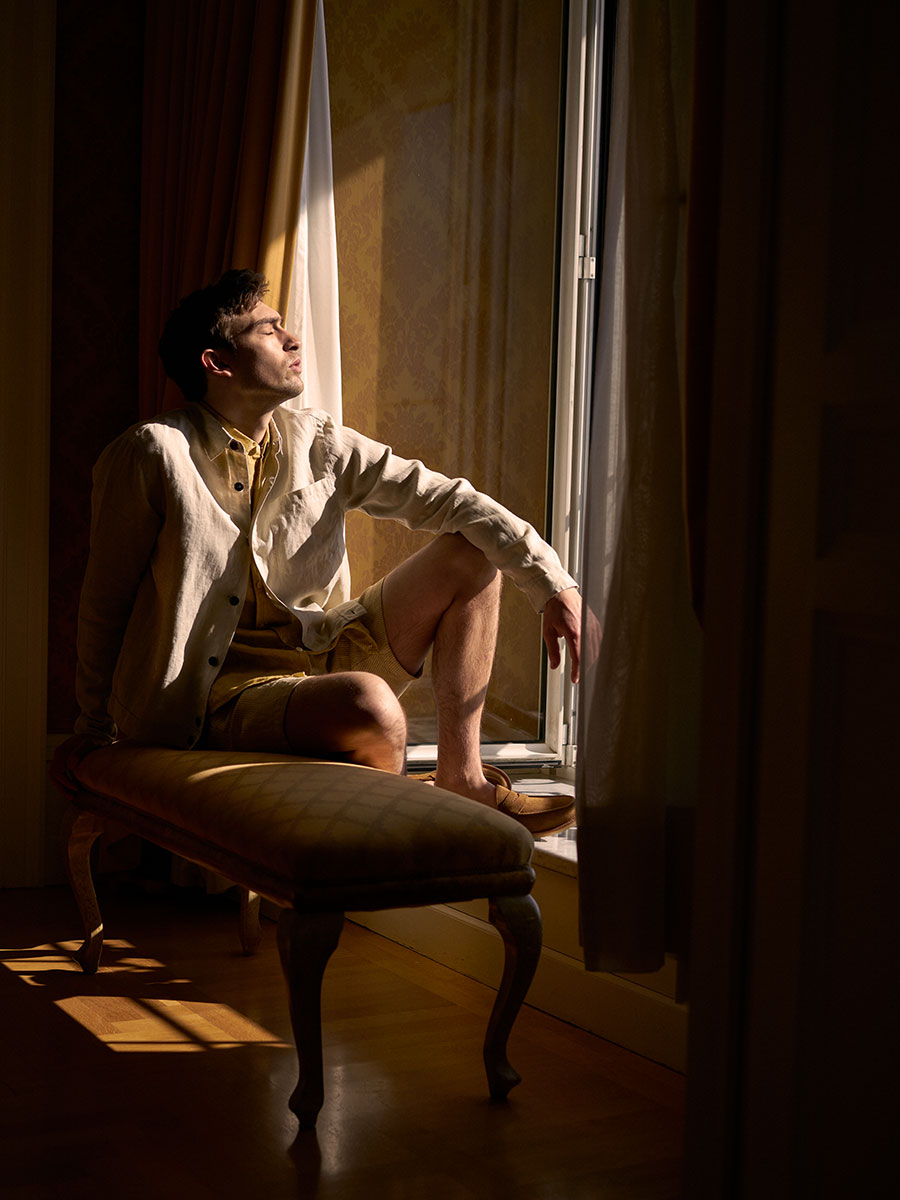 Oliver Spencer Summer 22 in Naples, look 16. Seated in a dark interior lit by a single window.